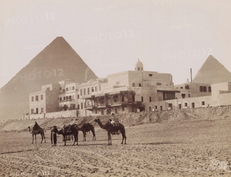 Mena House, View of the Mena House Hotel near Cairo, photograph, number: 'No 325', photographer: G. Lekegian & Co., Cairo (city), 1887 - 1900, Ottoman Empire, North Africa, paper, albumen print, height 211 mm x width 275 mm, hotel, hostelry, inn, pyramid (historical grave form), Mena House Hotel, A wide sandy foreground with visible hoof and foot tracks leads to a row of camels standing and being led by figures, some mounted and some standing beside the animals, the camels carrying saddles and decorative fringes; behind them a low retaining wall separates the sand from a terrace of multi-story stone and stucco buildings featuring balconies, shuttered windows, an ornate wooden veranda, arched openings, and a domed rooftop structure; in the background two large pyramidal stone structures rise against a pale sky.