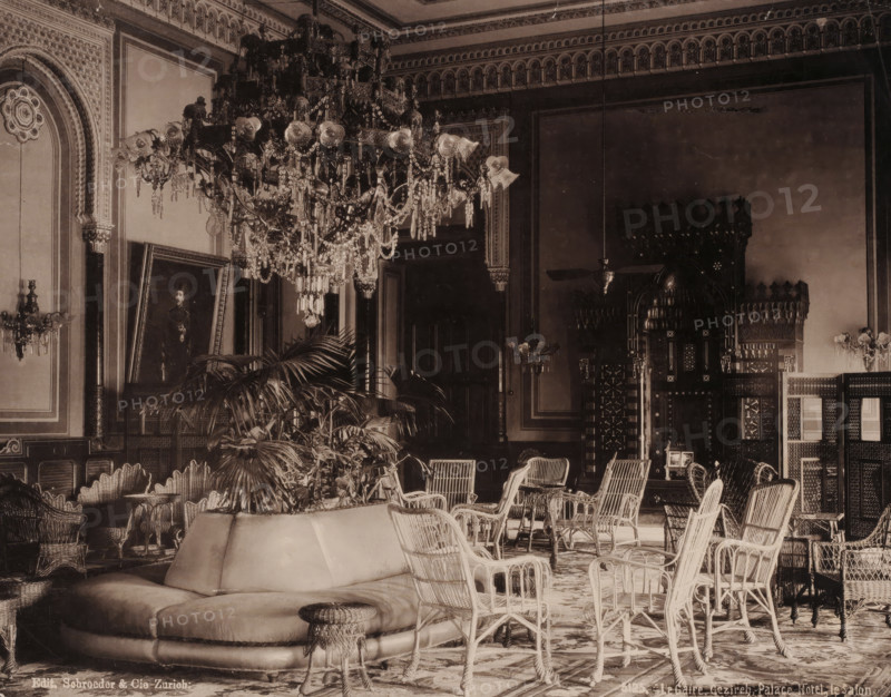 Le Caire, Gezireh, Palace Hotel, le Salon, Salon in the Gezirah Palace Hotel in Cairo, photomechanical print, number: '5125.', Gezirah, clichémaker: anonymous, publisher: Schröder & Co., Zurich (city), 1894 - 1895, photographer, Ottoman Empire, North Africa, Cairo (city), paper, collotype, photogravure, height 211 mm x width 269 mm, hotel, hostelry, inn, drawing-room, 'salon', Gezirah Palace, A large ornate chandelier with hanging crystals dominates the ceiling area above an elaborately decorated room with patterned cornices and framed wall panels; beneath it are wicker chairs and small tables arranged around a low upholstered circular sofa and a round woven stool, with potted palms and other plants placed near the seating; carved wooden screens and an intricately carved niche with geometric and arched details occupy the back wall, while framed artwork leans against a side wall and wall-mounted light fixtures with glass shades flank the room.