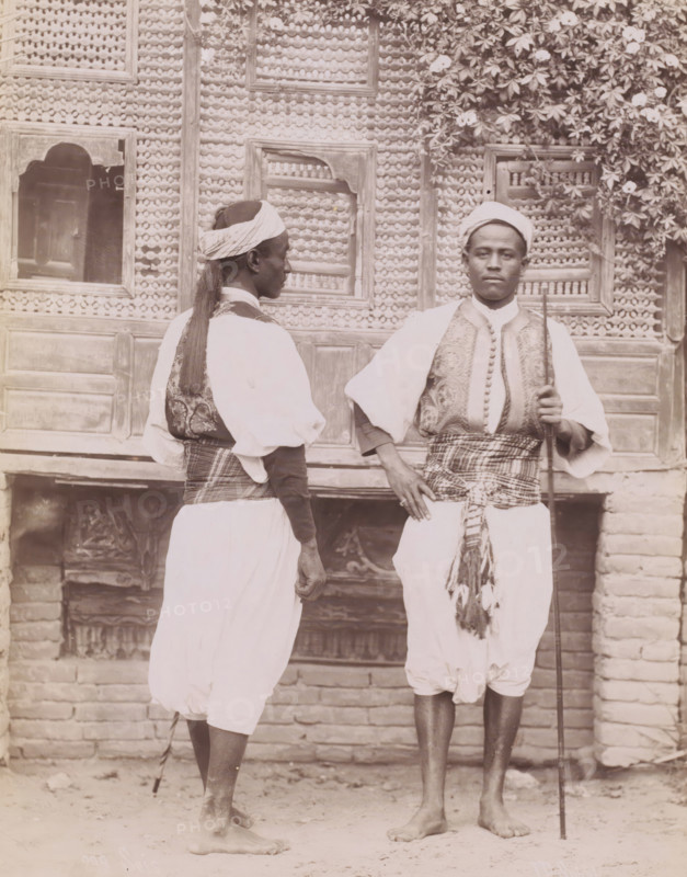 Double portrait of two unknown Egyptian soldiers (Séis), Séis, photograph, number: '289 ?', photographer: Jean Pascal Sébah, Egypt, 1888 - 1900, North Africa, paper, albumen print, height 270 mm x width 212 mm, the soldier, the soldier's life, Two men stand barefoot in front of a patterned wooden wall with carved panels and a flowering vine at the top right; one man faces slightly away showing a wrapped headscarf, long hair tied at the back, a patterned vest over a light shirt, and a cloth sash around his waist, while the other faces forward holding a long staff, wearing a wrapped head covering, an embroidered vest over a light shirt, a patterned sash tied at his waist with tassels, and loose light trousers.
