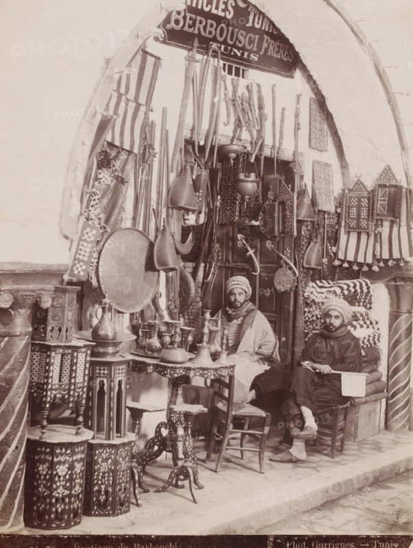 Barbouchi Frères in front of their shop in the Souk el-Trouk in the Medina of Tunis, Boutique de Barbouchi, photograph, number: 'N. 70', photographer: J. André Garrigues, Medina of Tunis, 1860 - 1900, North Africa, Tunis, paper, albumen print, height 258 mm x width 199 mm, shop, store, Souk el-Trouk, A storefront framed by a rounded arch displays hanging rifles and long wooden items, decorative shields, patterned textiles, and ornate metal lanterns and vessels; two men wearing wrapped head coverings and layered clothing sit among stacked folded fabrics and carved wooden tables and stools with inlaid designs, surrounded by small metal pitchers, trays, and intricately patterned boxes and panels.