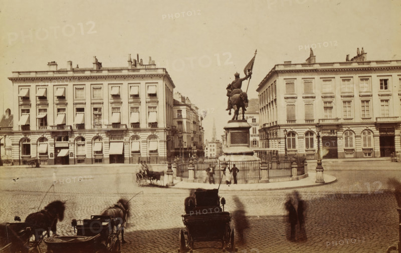 Koningsplein with the statue of Godfrey of Bouillon in Brussels, Belgium, photograph, Brussels, after sculpture by Louis Eugène Simonis, 1851 - 1900, Louis Eugène Simonis, photographer, after sculpture by, paper, albumen print, albumendruk, height 107 mm x width 164 mm, Person, Godfrey of Bouillon, monument, statue, square, circus, Koningsplein (Brussels), A wide cobblestone square with a central fenced pedestal supporting an equestrian statue of a rider holding a flag, flanked by multi-story classical buildings with rows of windows and awnings, chimneys and roof details; horse-drawn carriages and blurred figures in the foreground, lampposts near the statue, and a distant narrow street visible between the buildings leading to a spire.