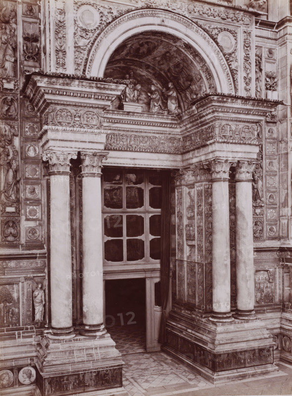 Porta della Chiesa, Portal of the monastery church Certosa di Pavia in Pavia, Italy, Certosa di Pavia, photograph, number: '13', inscription: 'Brogi (t)', number: '4621.', Pavia (city), publisher: attributed to Giacomo Brogi, Florence, 1856 - 1881, photographer, Tuscany, Lombardy, Attributed to, pasteboard, paper, albumen print, height 404 mm x width 306 mm, parts of church exterior and annexes: portal, A stone entrance framed by ornate carved columns and decorative pilasters with sculpted reliefs and medallions, an arched tympanum above filled with carved figures and painted panels, a deep recessed doorway with a paneled window above and a partially open curtain hanging to one side, carved capitals atop the columns, layered cornices with egg-and-dart and leaf motifs, patterned stone floor tiles at the threshold, and decorative friezes and sculpted figures integrated into the surrounding façade.
