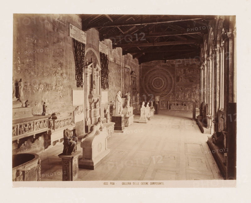 Sculpture gallery in the Camposanto in Pisa, Italy, Galleria delle Catene Camposanto, Pisa, photograph, number: '1022.', after sculpture by various manufacturers, 1851 - 1900, various manufacturers, photographer, after sculpture by, Tuscany, various makers, pasteboard, paper, albumen print, height 310 mm x width 383 mm, gallery of sculptures, Campo Santo (Pisa), A long colonnaded interior space with a high wooden beamed ceiling and a row of arched openings along the right side, stone floor tiles, and walls decorated with large circular and figurative wall paintings. Along the left wall there are sculpted tombs, ornate carved panels, and freestanding statues and busts on pedestals, with decorative reliefs and sculpted figures visible. Two large heavy chains hang beside a recessed architectural niche with an ornate frame and sculpted elements. Several sculpted reclining figures and seated figures rest atop tomb structures, and a freestanding column-like object stands near the far wall be...