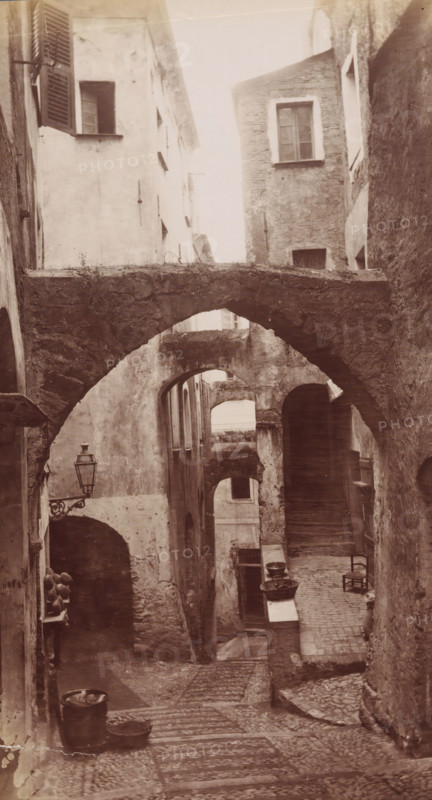 Alley in San Remo, San Remo, photograph, 1851 - 1900, photographer, paper, albumen print, albumendruk, height 271 mm x width 148 mm, street, A narrow, cobbled alleyway framed by tall, weathered stone and plaster buildings with multiple arched passageways and stone bridges crossing overhead; the walls show textured, worn surfaces and small windows with shutters and simple frames, one shutter partially open; a wrought iron street lamp projects from a wall near an arch; stone steps and uneven paving lead down the alley, where clay pots, basins, and a metal bucket sit on ledges and the ground; a simple wooden chair rests against a wall near a doorway and a small open arch reveals deeper recesses and stairways within the buildings.