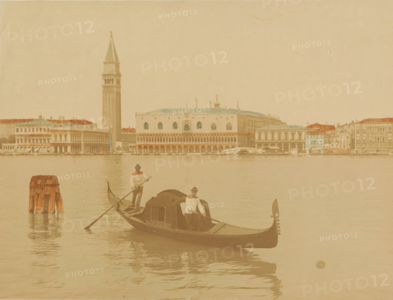 View of the Doge's Palace and the Campanile and in the foreground a gondola in Venice, Italy, photograph, Venice, 1851 - 1900, photographer, pasteboard, paper, albumen print, hand coloring, height 309 mm x width 399 mm, palace, gondola, parts of church exterior and annexes: campanile, Doge's Palace, Campanile (Venice), A wide waterfront scene with a tall bell tower and a large ornate building with arches and domes along the waterline, a row of multi-story buildings with varied roofs stretching across the background, a gondola in the foreground carrying a covered cabin and two standing oarsmen wearing hats and light shirts, wooden mooring posts emerging from the water to the left, calm reflective water filling the lower half of the image, and pale, muted tones throughout.