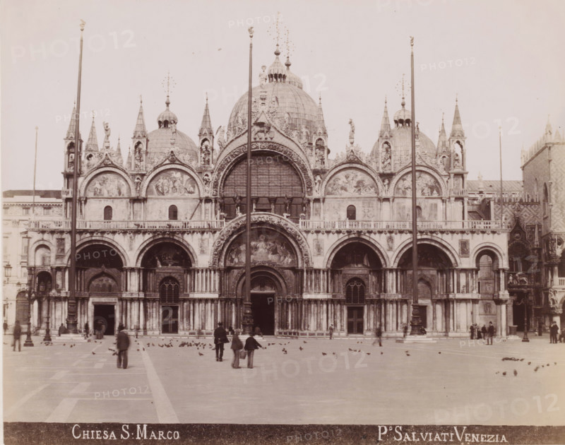 View of San Marco in Venice, Chiesa S. Marco, photograph, inscription: 'Venetie', photographer: Paolo Salviati, Venice, 1860 - 1870, pasteboard, paper, albumen print, albumendruk, height 199 mm x width 251 mm, height 275 mm x width 365 mm, square, circus, church (exterior), St. Mark's Basilica, St. Mark's Square, A wide facade of an ornately decorated cathedral fills the image, featuring multiple domes, pointed spires, arched doorways, and sculpted reliefs above the arches; a large central arch with detailed artwork sits above the main entrance, flanked by smaller arches and rows of columns; tall flagpoles rise in front of the building; a broad open plaza extends from the cathedral with scattered birds on the ground and several people walking or standing, some in groups and some alone; the overall image is monochrome with high contrast and an expansive sky above the building.