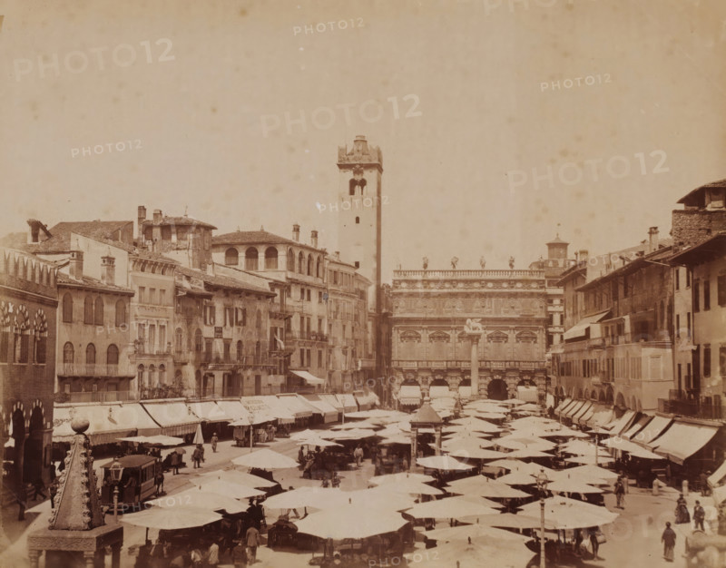 Market square in Verona, Italy, photograph, number: '13', inscription: 'Verona', inscription: 'Verona - Mercato ... Erbe ?', Verona, 1851 - 1900, photographer, pasteboard, paper, albumen print, albumendruk, height 304 mm x width 404 mm, market, square, circus, A crowded open market fills the foreground with rows of shaded stalls topped by large umbrellas and canopies, and people walking or standing among them; on the left a stone obelisk with carved details rises from a pedestal next to a covered tram and a small group of figures; surrounding the market are multi-story buildings with tiled roofs, balconies, arched windows, and various chimneys, and a tall square tower with an open belfry stands near the center background adjacent to an ornate façade decorated with sculpted figures and reliefs; the sky above is pale and largely empty.