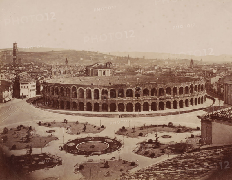 View of the Arena of Verona, Italy, photograph, inscription: 'Verona.', number: '13', Verona, 1851 - 1900, photographer, pasteboard, paper, albumen print, height 306 mm x width 405 mm, city-view in general, 'veduta', amphitheater, Arena of Verona, A wide sepia-toned cityscape centered on a large oval stone amphitheater with multiple tiers of arched openings and a curved exterior roadway, surrounded by low buildings and narrow streets; in front of the amphitheater is a landscaped plaza with circular flowerbeds, paved paths, benches, streetlamps, and small trees, with a few scattered people walking or sitting; beyond the amphitheater a dense cluster of historic buildings and towers stretches toward rolling hills in the background, and the sky above is pale and largely empty.