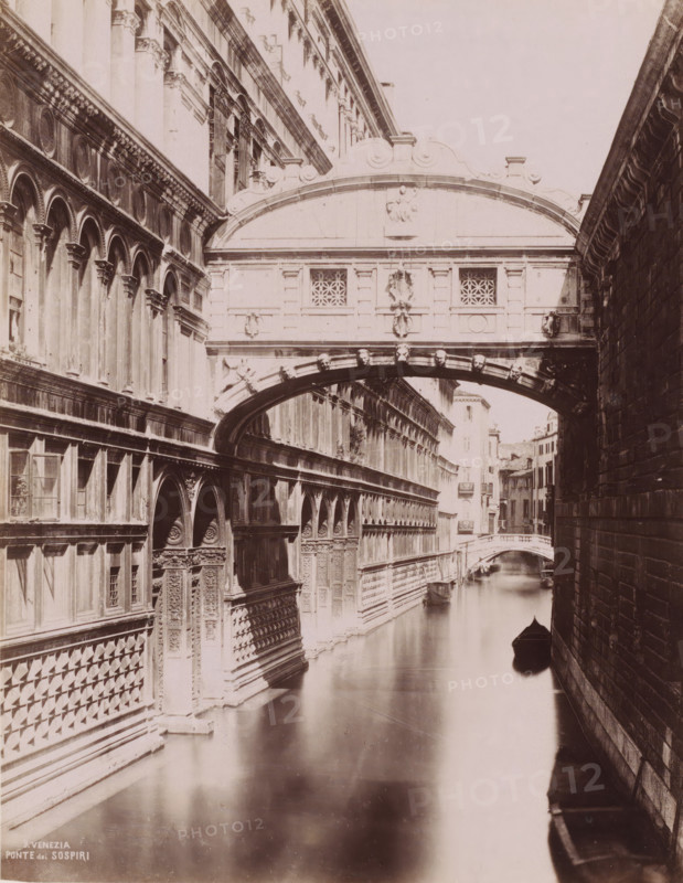 Ponte dei sospiri, Bridge of Sighs in Venice, Italy, Venezia, photograph, number: '9.', Venice, 1851 - 1900, photographer, pasteboard, paper, albumen print, albumendruk, height 369 mm x width 310 mm, canals, waters (in city), bridge, A narrow canal runs between two tall stone buildings with ornate facades, decorative arches, columns, carved reliefs, and patterned masonry; a covered arched bridge spans the canal with sculpted faces along its underside and carved panels and lattice windows on its exterior; a small boat with a pointed bow floats on the calm reflective water near the shadowed side of the right-hand wall; a distant arched bridge and more buildings line the canal further back under soft lighting.