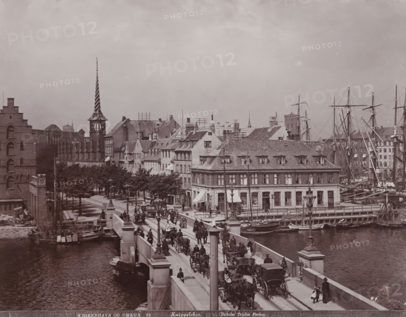 Knippelsbro, View of the Knippelsbro in Copenhagen, Kjøbenhavn og Omegn, photograph, number: '52.', Copenhagen (city), publisher: Vilhelm Trydes, Denmark, 1880 - 1910, photographer, paper, pasteboard, baryta paper, daylight collodion silver print, height 222 mm x width 283 mm, height 349 mm x width 455 mm, bridge in city across river, canal, A wide city scene centered on a stone bridge crowded with people, horse-drawn carriages, and pedestrians, flanked by river water with small boats moored along the quay, a row of multi-story buildings with tiled roofs and dormer windows lining the waterfront, tall masts and rigging of sailing ships rising behind the buildings, a narrow street with trees and lampposts leading into the scene, a distinctive tower with a twisted spire visible among the rooftop silhouettes, and numerous architectural details such as chimneys, windows, shop awnings, and street lamps.