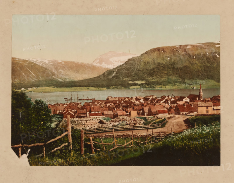 Village view in Norway, photomechanical print, Norway, cliché maker: anonymous, 1860 - 1900, photographer, pasteboard, paper, albumen print, color lithography, albumen printing with color lithography, height 165 mm x width 228 mm, height 349 mm x width 457 mm, city-view in general, 'veduta', A panoramic view shows a village with clustered houses topped by reddish-brown roofs and light-colored walls, a church with a tall steeple near the water, a shoreline with boats and masts visible on the calm water, rolling green fields and fenced pastures in the foreground with a dirt path leading toward the village, dense trees and shrubs near the front left, and distant mountains with patches of snow under a pale sky.
