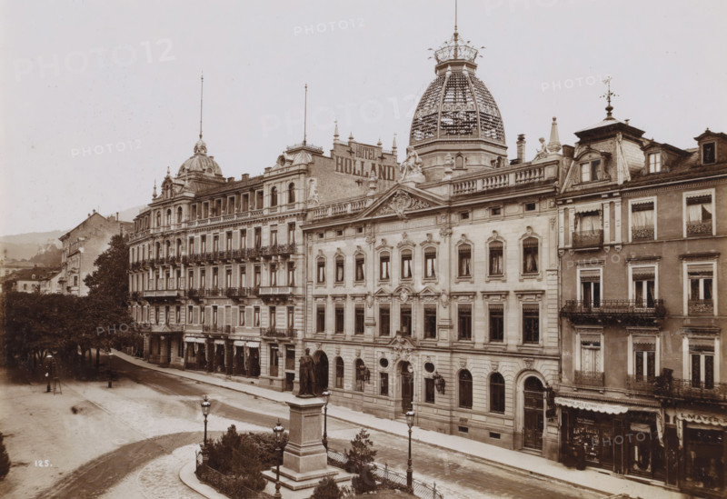Street view in German city (former title), View of Leopoldsplatz in Baden-Baden, photograph, In the middle the post office ('Kaiserliches Postamt') and next to it Hotel Holland., check stamp: '1903, Gust. Salzer, B. Baden', number: '125', photographer: Gustav Salzer, Baden-Baden, 1903, Baden-Württemberg, paper, baryta paper, daylight collodion silver print, height 115 mm x width 164 mm, square, circus, hotel, hostelry, inn, post-office, A wide street scene shows a row of ornate multi-story stone and brick buildings with decorative facades, balconies with wrought iron railings, and numerous tall windows with curtains; a large domed tower with an intricate lattice structure rises above one building, flanked by smaller domes and spires topped with finials; sculptural figures and ornamental stonework decorate the rooflines and pediments; a broad sidewalk runs along the buildings and a tree-lined area with mature trees and shrubs sits opposite; a stone monument with a statue atop a tall ped...