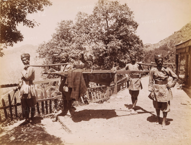 Four Indian men in unform life a palanquin, India, photograph, signature: 'SH Dagg', inscription: '£1', inscription: 'Asia', photographer: Sidney Herbert Dagg, British India, 1890 - 1910, pasteboard, paper, albumen print, height 212 mm x width 273 mm, height 227 mm x width 292 mm, (carrying a person on a) litter, stretcher, Four men wearing turbans and tunics stand on a dirt path beside a wooden fence and railing, two of them holding a long wooden pole across their shoulders that supports a stretcher-like frame draped with a cloth, while a large tree with dense foliage fills the background and a hillside and a simple building with a slanted roof appear to the right.