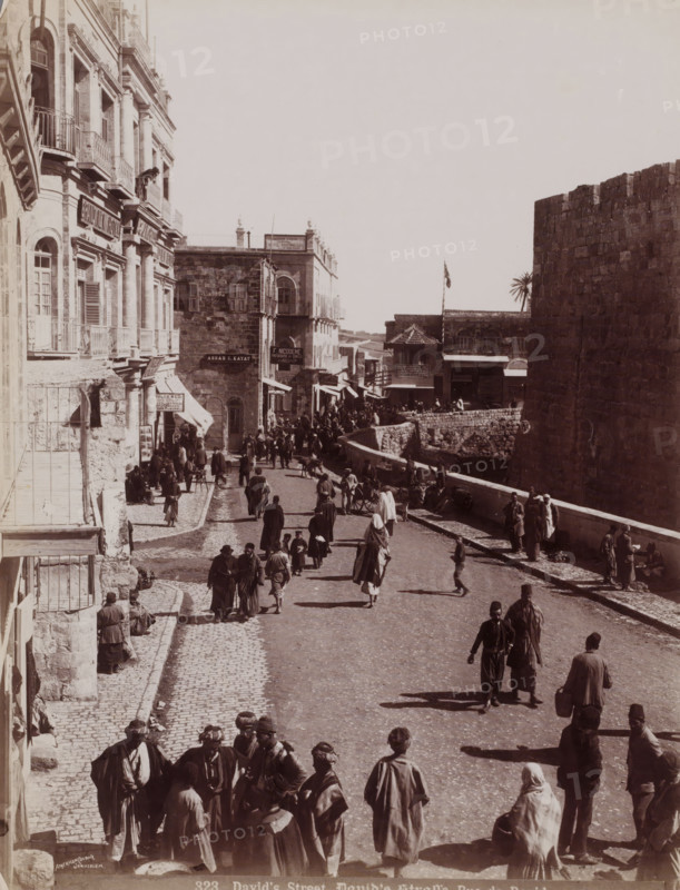 David's Street, David's Straße, Rue de David, View of David Street in Jerusalem, in the distance the photo shop of F. Nicodeme, photograph, number: '323', photographer: American Colony, Jerusalem, 1898 - 1910, paper, albumen print, height 227 mm x width 215 mm, street, A wide street scene with a stone-paved road flanked by multi-story buildings on the left and a high stone wall on the right, populated by many people walking, standing, and sitting wearing long coats, robes, and head coverings; balconies and shuttered windows project from the buildings, and awnings and small shopfronts line the left side while a low stone parapet runs along the right beside the wall; a flag flies near the center background above a building with a tiled roof, and carts and groups of people gather further down the street; shadows fall across the cobblestones and the overall image is monochrome.