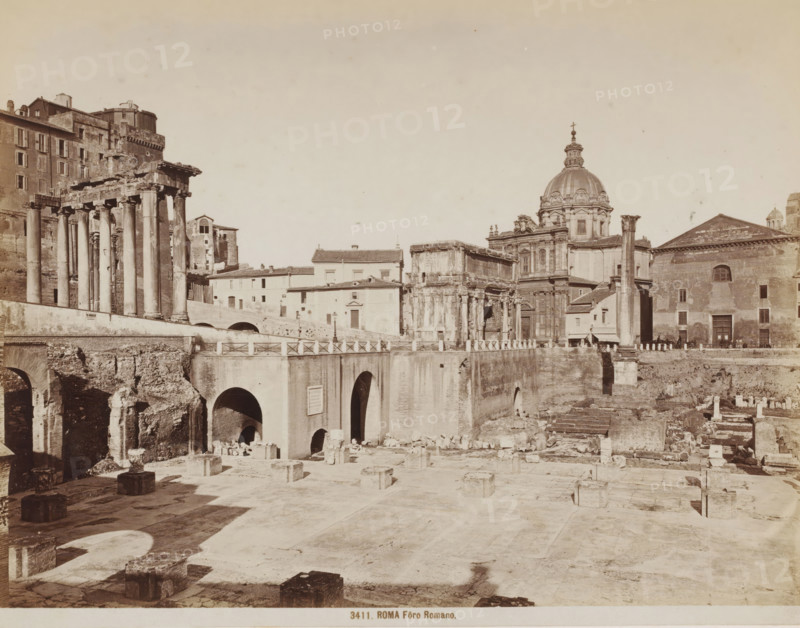 View of the Roman Forum in Rome, Italy, Fôro Romano, Roma, photograph, number: '53-', number: '3411.', number: '9', photographer: attributed to Giacomo Brogi, Rome, 1864 - 1881, Attributed to, pasteboard, paper, albumen print, height 276 mm x width 374 mm, landscape with ruins, excavation, archaeology, Roman Forum, A wide sepia-toned scene of ancient ruins and classical buildings with a foreground of a flat stone plaza scattered with square stone bases and low walls, a middle ground dominated by a large stone bridge with multiple arched openings and a balustrade, and to the left a partially preserved colonnade of tall columns supporting a broken entablature beside multi-story masonry buildings; to the right a cluster of ornate stone structures including a domed church with decorative architectural details and a façade with pilasters, and adjacent rectangular and temple-like buildings with single standing columns and arched doorways, all set beneath a pale sky.