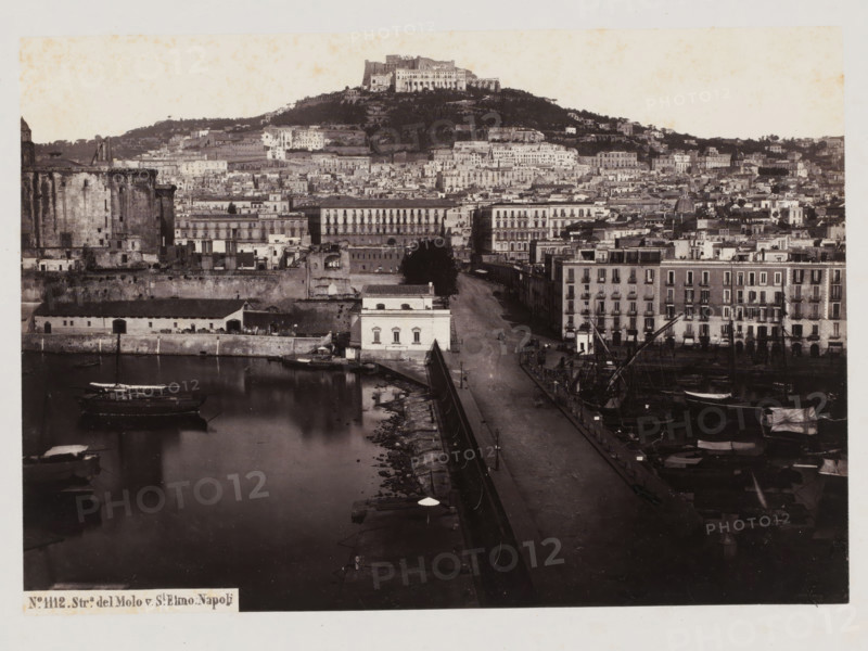 View of the Sant'Elmo castle on Mount Vomero in Naples, Italy, Strada del Molo v. St. Elmo, Napoli, photograph, number: 'No. 1112.', number: '22', inscription: 'Naples.', address: 'GIORGIO SOMMER, STUDIO MONTE DI DIO 4., MAGAZZINO S. CATERINA 5., NAPOLI', photographer: Giorgio Sommer, Naples, 1890 - 1910, Giorgio Sommer, photographer, pasteboard, paper, albumen print, height 307 mm x width 377 mm, city-view in general, 'veduta', castle, Castel Sant'Elmo, A black and white panoramic view of a waterfront and an urban hillside showing a harbor with moored boats and masts, a long quay lined with lamp posts and a paved road, waterfront buildings with multiple stories and balconies, a lighter colored rectangular building close to the water, a large round stone fortification on the left, dense rows of varied buildings climbing the hillside, terraces and trees on the slope, and a prominent castle-like structure perched atop the hill.