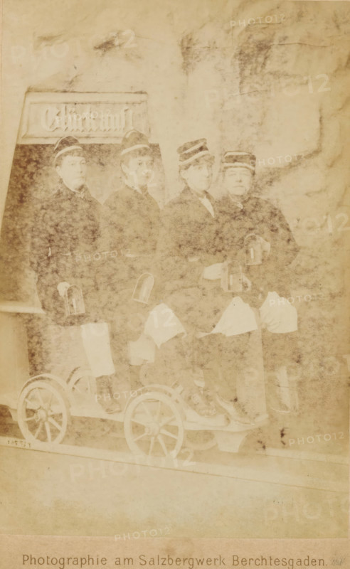 Group portrait of four women wearing caps, sitting on a carriage on a rail, cabinet photograph, name: 'P. Ney Atelier für Photographie am Königl. Bayerischen Salzbergwerk Berchtesgaden, ', photographer: Paul Ney, Berchtesgaden, c. 1870 - c. 1900, Bavaria, paper, pasteboard, albumen print, height 148 mm x width 97 mm, adult woman, railway, train, A faded sepia photograph showing four people seated side by side on a small wooden cart with visible spoked wheels, each person wearing a dark jacket and a cap with a light band, lighter cloth draped across their laps, and small hand lanterns held or attached near their hands, with a rectangular signboard partially visible behind them and a mottled background surrounding the group.