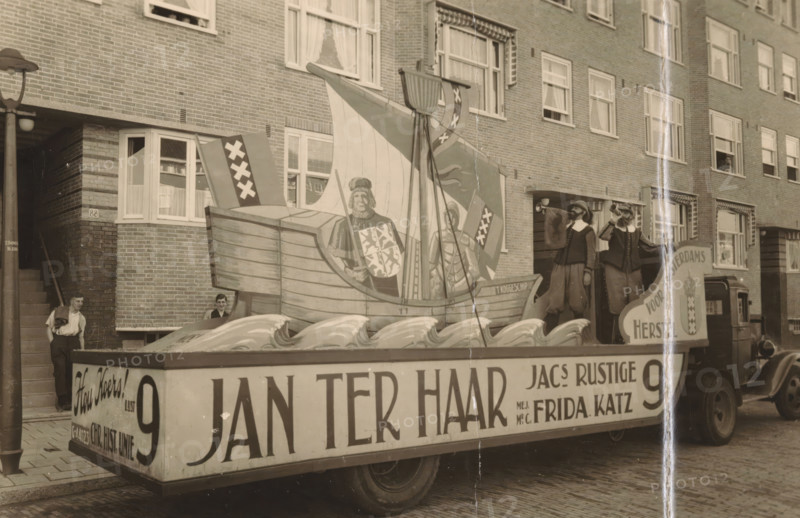 Decorated float of the political party List 9 during a parade in Amsterdam, photograph, postcard, annotation: 'Amsterdam, 1930s', photographer: T en M, Amsterdam, 1920 - 1940, paper, gelatin silver print, height 89 mm x width 139 mm, political parties, public festivities (parade, pageant, cavalcade, festive activities), A street scene showing a large parade float on a flatbed truck in front of a brick apartment building, the float featuring a painted wooden ship with a tall sail, a painted figure on the ship holding a shield, decorative stylized waves along the float base, two costumed men standing on the float blowing horns, a man leaning against a doorway at the building entrance and another person partially visible near a window behind the float, multi-pane windows with curtains across the building facade, a lamppost at the left edge, and cobblestone pavement beneath the truck.