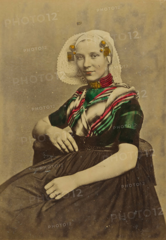 Portrait of an unknown woman in traditional costume of Goes, Zeeland, cabinet photograph, annotation: 'Goes, R..., Costumes des Pays Bas., Publié par A. Jager, Damrak 81, Amsterdam', annotation: 'Zeeland traditional costume.', number: '40', photographer: Andries Jager (possibly), Amsterdam, 1860 - 1890, paper, pasteboard, albumen print, hand coloring, height 156 mm x width 115 mm, folk costume, regional costume, historical persons not known by name - BB - woman, A seated young woman wearing a lace headpiece with rounded edges and decorative adornments at the temples, a striped shawl with bright narrow bands draped over a dark short-sleeved bodice, and a high necklace of small beads; she has a composed facial expression and is looking toward the viewer, one arm resting on an armrest and the other hand placed on her skirt, with rings visible on fingers and a pleated dark skirt extending across her lap.
