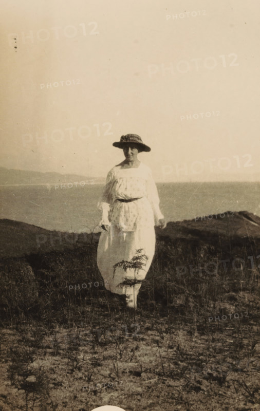 Portrait of an unknown woman near a coast, photograph, inscription: 'Voor Kees, from aunt Cor', 1919-09-28 - 1929-09-28, photographer, paper, baryta paper, gelatin silver print, developing gelatin silver print, height 135 mm x width 86 mm, anonymous historical persons in a double portrait - BB - woman, A sepia-toned photograph of a person standing outdoors on grassy, sandy ground near a body of water, wearing a light-colored ankle-length dress with a patterned apron or overdress and a wide-brimmed hat with a decorative band, the person facing the camera with hands relaxed at their sides, low hills visible across the water in the background and sparse vegetation in the foreground.