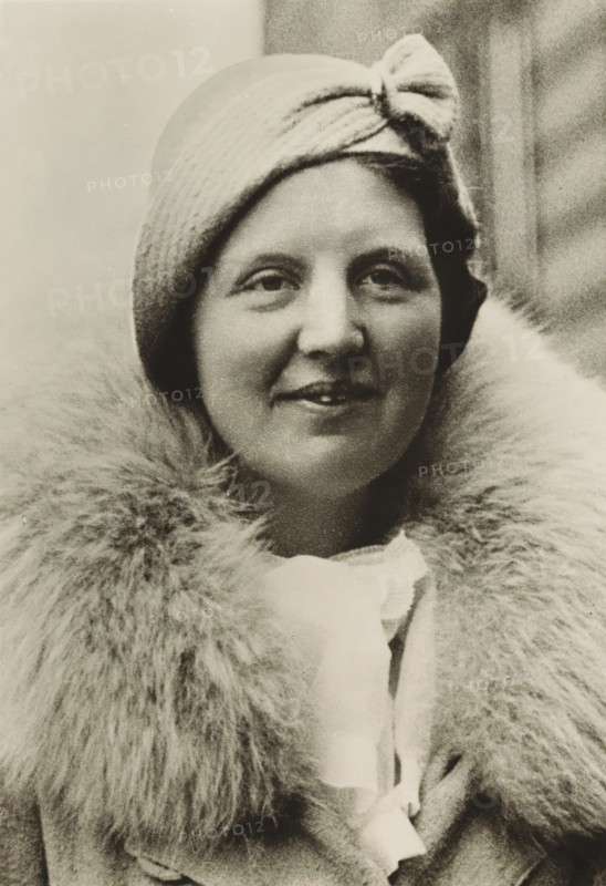Portrait of Princess Juliana, photograph, picture postcard, London, publisher: Have, W. ten, Amsterdam, 1934, photographer, paper, gelatin silver print, developing gelatin silver print, height 153 mm x width 105 mm, Person, Juliana of the Netherlands, historical persons, crown princess, A close-up portrait of a woman wearing a cloche-style hat with a knot detail at the front and a large, fluffy fur collar around her neck; she has a slight smile, visible eyes and lips, and smooth facial features, with the background showing a softly textured, out-of-focus surface.