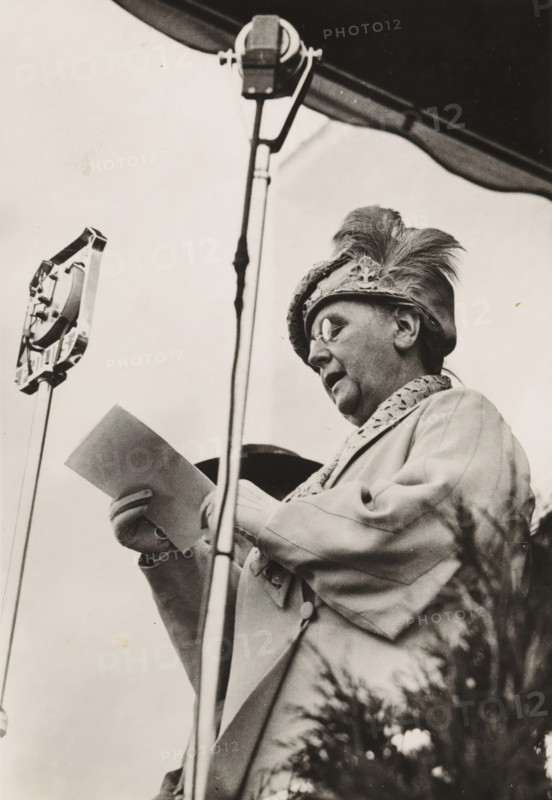 HM the Queen opens the World Jamboree July 31, 1937, photograph, postcard, Vogelenzang (North Holland), publisher: Have, W. ten, Amsterdam, 1937-07-31, photographer, paper, gelatin silver print, height 151 mm x width 105 mm, Person, Wilhelmina of the Netherlands, queen, A black-and-white image of an older person wearing round glasses and a feathered hat, shown from a low angle behind a microphone stand, holding a sheet of paper and dressed in a coat with a patterned collar.