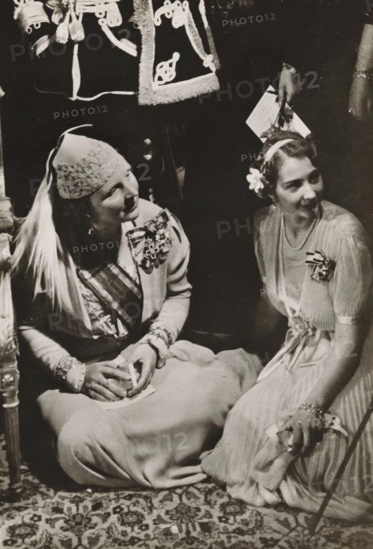 HRH Princess Juliana with Princess Cecilie, youngest sister of Prince Louis Ferdinand, as a guest at the wedding between Grand Duchess Kyra and Prince Louis Ferdinand, photograph, postcard, Doorn, publisher: Have, W. ten, Amsterdam, 1938-05-04, photographer, printer (person), paper, gelatin silver print, gelatin silver developing print, height 152 mm x width 105 mm, Person, Juliana of the Netherlands, prince, A black-and-white photograph showing two seated women dressed in formal attire, both wearing decorative headpieces and floral corsages; the woman on the left wears a textured cap with a draped veil, a jacket with ornate medals and brooches, bracelets, and holds a small folded item in her hands, while the woman on the right wears a headband with a flower, a string of pearls, a light-colored pleated dress with a ribbon at the waist, and rests one hand on a cane; patterned carpet is visible beneath them and a portion of another person's decorated garment appears at the top of the fra...