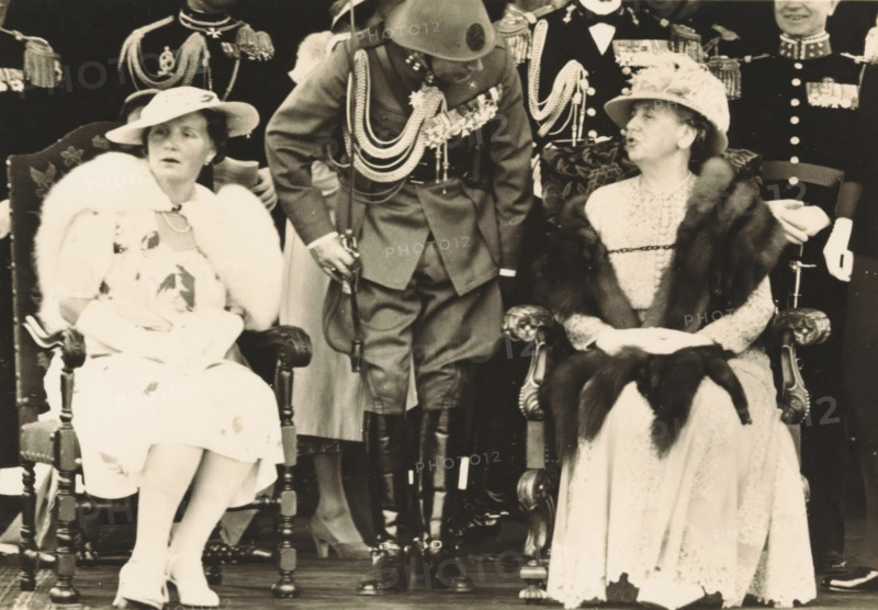 Army Day in The Hague, photograph, picture postcard, The Hague, publisher: Have, W. ten, Amsterdam, 1938-08-29, photographer, printer (person), paper, gelatin silver print, developing gelatin silver print, height 105 mm x width 150 mm, Person, Juliana of the Netherlands, Wilhelmina of the Netherlands, queen, crown princess, A black-and-white photograph showing two seated women in ornate chairs and a uniformed man standing between them; the woman on the left wears a light-colored dress with a fur stole, a wide-brimmed hat, and white gloves, her legs crossed and shoes visible, while the woman on the right wears a patterned dress with a fur wrap, a decorated hat, and gloves, her hands clasped in her lap; the man between them wears a military-style uniform with decorative cords across his chest, a helmet, high boots, and holds a sword; behind them are other uniformed figures with visible epaulettes and medals.