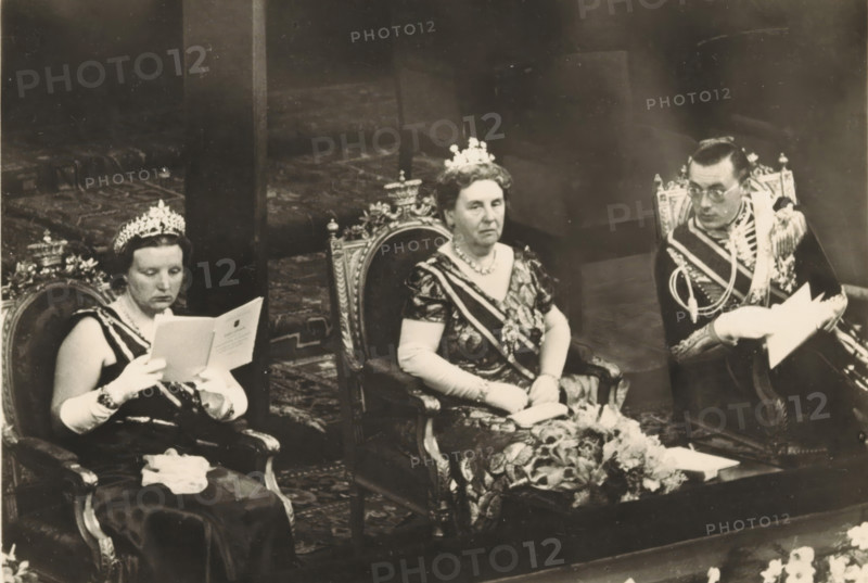 A few days before the 40th anniversary of the reign of HM the Queen. Evening of Honour in the Building for Arts and Sciences in The Hague, photograph, postcard, The Hague, publisher: Have, W. ten, Amsterdam, 1938-08-31, photographer, printer (person), paper, gelatin silver print, height 105 mm x width 150 mm, Person, Bernhard (Prince of the Netherlands), Juliana of the Netherlands, Wilhelmina of the Netherlands, queen, prince, crown princess, Building for Arts and Sciences (The Hague), Three seated people wear formal attire and crowns while sitting on ornate chairs; the left person reads from a folded paper, wearing long gloves and a dark dress with a sash, the central person faces forward with a patterned dress, gloves, a sash, and a necklace, and the right person wears a decorated uniform with braided cords and holds papers. The chairs have carved, decorative backs, and a patterned carpet is visible beneath them. There is a floral arrangement on a low table in front of the central pe...