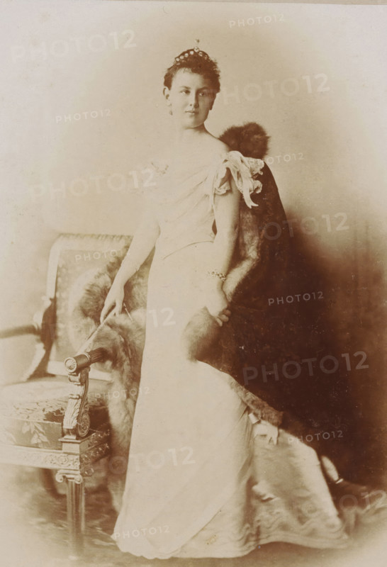 Portrait of Queen Wilhelmina, cabinet photograph, stamp: 'GEBR. DOUWES, ATELIERS, Warmoesstraat 73., AMSTERDAM.', 1896 - 1899, photographer, paper, pasteboard, daylight collodion silver print, height 166 mm x width 107 mm, Person, Wilhelmina of the Netherlands, historical persons, queen, A sepia-toned portrait of a woman standing beside an ornate upholstered chair, wearing a light-colored off-the-shoulder gown with gathered sleeves and a long skirt, a small tiara in her hair, and a fur-trimmed cloak draped behind her; one hand rests lightly on the chair while the other holds the fur, and a bracelet is visible on her wrist.