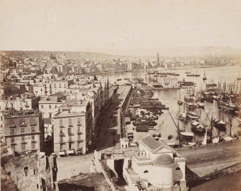Port et rivière Carmine, View of the port of Naples and the river, Italy, Naples, photograph, inscription: '11.', number: '12', number: 'No. 4005.', inscription: '(+) Sommer', photographer: attributed to Giorgio Sommer, 1857 - 1914, Attributed to, pasteboard, paper, albumen print, albumendruk, height 305 mm x width 405 mm, harbour, city-view in general, 'veduta', A wide aerial view of a coastal city with rows of multi-story buildings lining a long waterfront street, numerous small boats and larger sailing vessels clustered along piers and floating in the harbor, a prominent domed building and other rooftop structures in the foreground, narrow streets between closely set buildings casting long shadows, a tower rising above the distant cityscape, distant low hills on the horizon, and calm water reflecting masts and hulls.