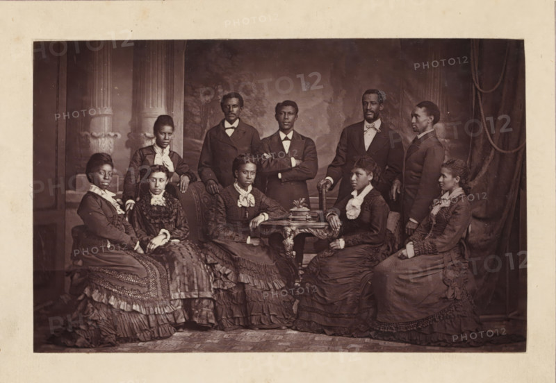 Group portrait of the Jubilee Singers, page, photomechanical print, cliché maker: anonymous, 1872 - in or before 1877, photographer, paper, Woodburytype, height 82 mm x width 130 mm, Print opposite title page., Person, Edmund W. Watkins, Hinton Alexander, Mabel Lewis, Frederick J. Loudin, Thomas Rutling, Maggie Carnes, Maggie Porter, Jennie Jackson, Ella Sheppard, America W. Robinson, historical persons, female singer, male singer, A sepia-toned studio portrait showing a group of adults arranged around an ornate wooden table and upholstered chairs, with decorative columns and drapery in the painted backdrop. The group includes men standing behind and women seated in front, all wearing formal Victorian-era clothing such as dark suits with bow ties for the men and long, layered dresses with ruffles, lace collars, and decorative trims for the women. Several women have their hands resting on the table or on their laps, and one man stands with his arms crossed. Facial expressions are neutra...