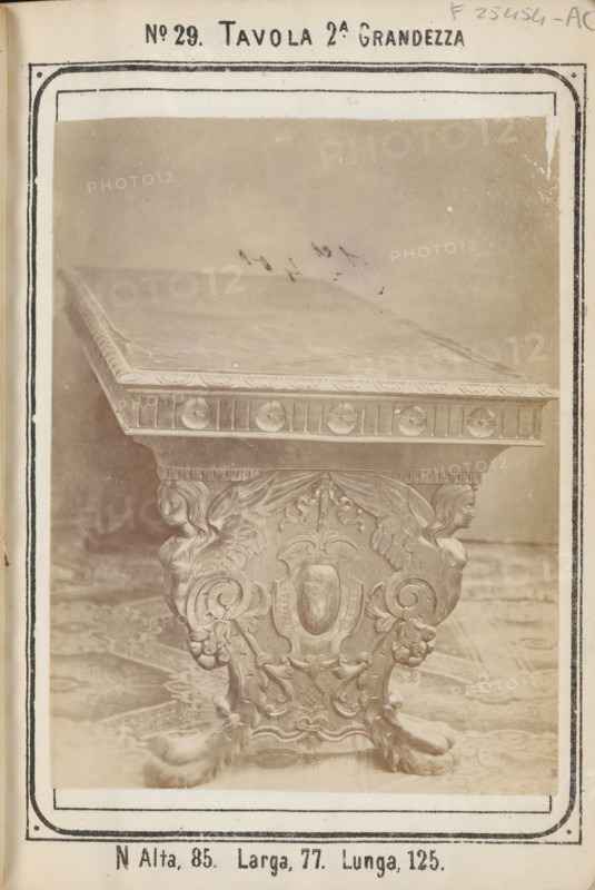 Table with decorated legs, No 29 Tavola, photograph, page, inscription: '2A. Grandezza, N Alta, 85. Larga, 77. Lunga, 125.', inscription: 'Ies. 70', c. 1878 - c. 1885, photographer, paper, albumen print, height 126 mm x width 96 mm, Photo on unnumbered page., table, A sepia-toned photograph of an ornately carved table top and its pedestal, showing a flat rectangular surface with a decorative edge and a frieze with circular motifs, supported by a richly carved central leg featuring symmetrical scrollwork, a shield-shaped cartouche, floral elements, and two carved female heads with wings, resting on claw-like feet and set on a patterned floor, all framed by a printed border and page margins with printed headings and marginal markings.