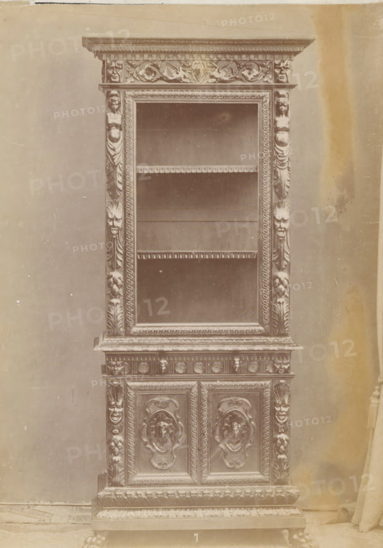 Kast, No 7 Mobile, photograph, page, inscription: 'Alto, 218. Largo, 92. Fondo, 50.', inscription: 'Ia. 130', c. 1878 - c. 1885, photographer, paper, albumen print, height 135 mm x width 96 mm, Photo on unnumbered page, cupboard, A tall, ornate wooden cabinet with carved decorative molding and relief panels is shown against a plain backdrop, the upper section featuring an open display area with shelves framed by carved borders and fluted pilasters adorned with sculpted faces and floral motifs, the lower section composed of paneled doors with raised, circular medallion carvings and intricate scrollwork, the base resting on carved feet and embellished with repeated ornamental patterns, the surface showing a uniform, aged tone and slight surface blemishes.