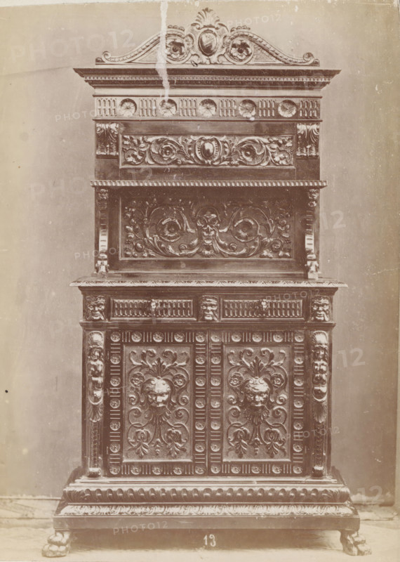 Decorated sideboard, No 13 Buffet, photograph, page, inscription: 'Ies. 160', inscription: 'Alto, 215. Largo, 111. Fondo, 49.', c. 1878 - c. 1885, photographer, paper, albumen print, height 131 mm x width 96 mm, Photo on unnumbered page, ornamental cupboard, A tall ornate wooden cabinet fills the frame, showing its upper decorative crest, carved panels, and base. The top features a curved carved pediment with floral and shell-like motifs and a central oval medallion, with a vertical lighter streak running down from near the crest. Below that is a horizontal frieze with circular rosette carvings and fluted details, followed by a recessed panel with intricately carved scrolling foliage and a central cartouche. The middle section has a projecting shelf with a row of small repeating rectangular dentils along its edge and a large carved panel beneath depicting swirling acanthus leaves and a stylized face at the center. Flanking columns with carved vertical ridges and small sculpted faces su...