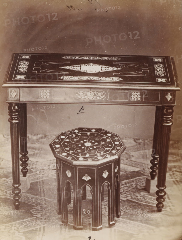 Table and stool with inlay, No 20 Tavola, Tambouret, photograph, page, number: '1., 2.', inscription: 'No 1. Ies. 180, ' 2 ' 45.', inscription: 'Alta, 76. Larga, 88. Fonda, 50., Alto 40. Largo 37.', c. 1878 - c. 1885, photographer, paper, albumen print, height 127 mm x width 95 mm, Photo on unnumbered page., table, stool, A close view of a wooden table with carved and inlaid decorative patterns on its flat top and apron, supported by turned, spiral legs, with a small octagonal wooden stool placed beneath it that has a patterned top and arched cutouts along its sides; a textured floor covering is visible under the furniture and the background above the table is plain and slightly mottled.