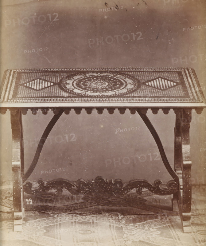 No 22 Tavola, Table with marquetry, photograph, page, inscription: 'Alta, 87. Larga, 122, Fonda 69.', inscription: 'Ies 200', c. 1878 - c. 1885, photographer, paper, albumen print, height 126 mm x width 109 mm, Photo on unnumbered page., table, A rectangular table with a patterned top featuring a central circular motif and two diamond-shaped checkerboard designs, supported by carved wooden legs and curved braces, with a decorative apron beneath the tabletop and a carved stretcher connecting the legs, all shown against a plain background and a rug partially visible on the floor.