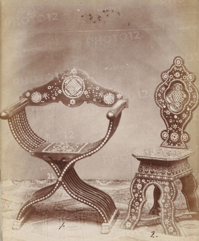 No 24 Poltrona, Sedia, Two types of chairs, photograph, page, inscription: 'No 1. Ies. 60., ' 2. 45', number: '1. 2.', inscription: 'Alta, 94. Larga, 70. Fonda, 36., ' 1m. '32. '35.', c. 1878 - c. 1885, photographer, paper, albumen print, height 127 mm x width 104 mm, Photo on unnumbered page., chair, A sepia-toned photograph showing two ornately decorated wooden chairs positioned on a patterned floor against a plain backdrop; the chair on the left has curved armrests, a decorative crest on the back with circular dot patterns, a curved slatted seat, and legs formed by interlacing curved slats with rows of round dot embellishments; the chair on the right features a tall, carved back with layered ornate shapes filled with dot motifs, a square seat with a patterned surface, and carved legs decorated with similar circular dot designs.