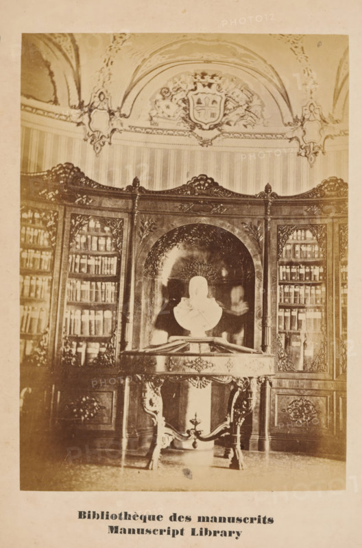 Bibliothèque des manuscrits, Manuscript Library, Library with manuscripts in the monastery at San Lazzaro degli Armeni, Venice, photograph, page, number: '9', c. 1874 - in or before 1879, photographer, paper, albumen print, height 125 mm x width 89 mm, Photo on unnumbered page opposite page 9., abbey, monastery, convent, Roman Catholic church, public library, San Lazzaro degli Armeni, A sepia-toned photograph of an ornate interior showing a carved wooden bookcase with glass-fronted shelves filled with books flanking a central arched niche, above which is decorative molding and a crest within a curved ceiling panel; in the niche a white bust sits on a small pedestal placed on an elaborately carved table with curved legs and decorative apron, and patterned wallpaper and ornate ceiling ornamentation are visible around the upper walls.