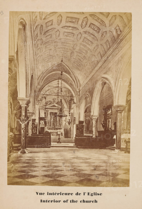 Photo reproduction of a drawing of the interior of the church of San Lazzaro degli Armeni, Venice, Vue intérieure de l'Eglise, Interior of the church, photograph, reproduction, page, number: '19B', after drawing by anonymous, c. 1874 - in or before 1879, photographer, after drawing by, paper, albumen print, height 128 mm x width 89 mm, Photo on unnumbered page opposite page 19., interior of church, San Lazzaro degli Armeni, A sepia-toned photograph of a large church interior showing a patterned floor with alternating light and dark tiles leading toward an elevated altar area framed by arches and columns, a vaulted ceiling decorated with recessed geometric panels, hanging chandeliers descending from the ceiling, ornate altar furnishings and statues positioned behind and around the altar, tall archways and columns lining the nave, a crucifix mounted on a stand near the foreground at one side, wooden pews and decorative architectural details along the side walls, and a wide aisle running ...