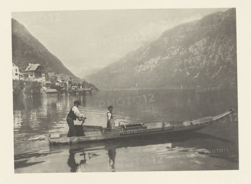 Boat on the Hallstätter See, Lac de Hallstatt, photomechanical print, photographer: Albert baron de Rothschild, Hallstatt, cliché maker: Jacob Blechinger, printer (person): Charles Wittmann, Paris, c. 1891 - in or before 1896, printer (person), Upper Austria, paper, photoaquatint, height 112 mm x width 156 mm, lake, mountains, rowing facing forward, pushing, Hallstätter See, A wide, calm lake bordered by steep, forested mountains fills the scene, with a cluster of lakeside buildings and boathouses along the shore at the left. Two small wooden boats float near the foreground, one with a person standing and another person leaning forward holding an oar; their reflections appear in the water. The overall image is monochrome with soft contrasts and a misty atmosphere over the distant mountains.