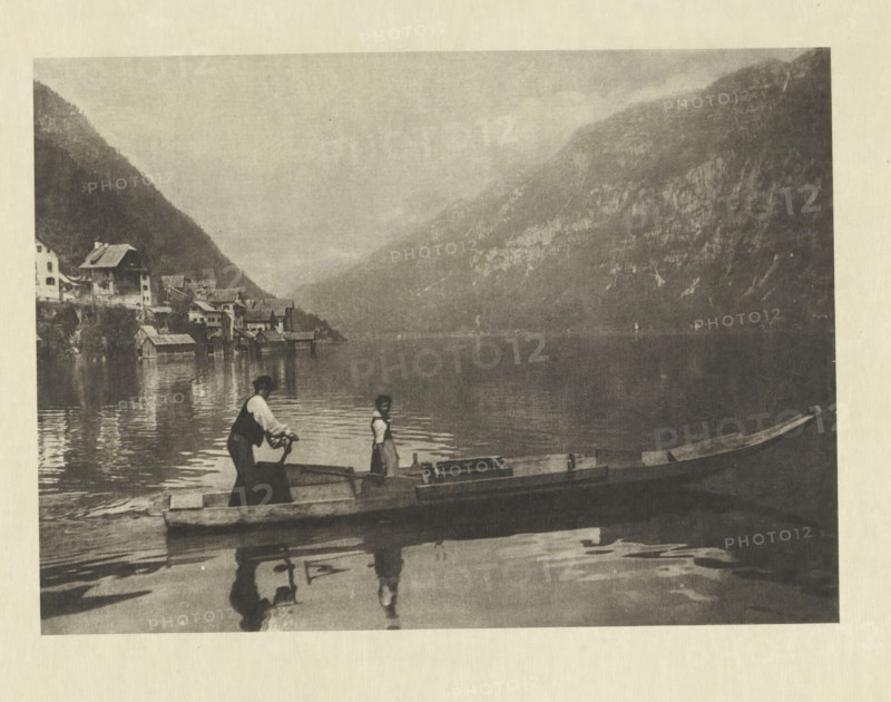 Lac de Hallstatt, Boat on the Hallstätter See, photomechanical print, photographer: Albert baron de Rothschild, Hallstatt, cliché maker: Jacob Blechinger, printer (person): Charles Wittmann, Paris, c. 1891 - in or before 1896, printer (person), Upper Austria, paper, photoaquatint, height 113 mm x width 157 mm, lake, mountains, rowing facing forward, pushing, Hallstätter See, A sepia-toned scene showing a calm lake bordered by steep mountains and a cluster of waterside buildings, with small wooden boats in the foreground and two people standing in one boat, their reflections visible in the water.