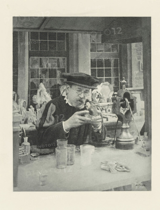 Man engraving on glass, Le Graveur sur fout, photomechanical print, photographer: Henri Colon, cliché maker: Fillon & Heuse, printer (person): Charles Wittmann, Paris, c. 1891 - in or before 1896, printer (person), paper, photoaquatint, height 179 mm x width 141 mm, handicrafts, glass, An older man wearing a dark beret and a dark garment with a light ruffled collar sits at a workbench, holding a small rounded object in his gloved hands and peering at it through round spectacles; behind him are window panes and shelves holding sculpted busts and other objects, while on the bench in front of him are glass containers, a cloth, a tall metal device with a crank and pulley, and various small tools and bottles.