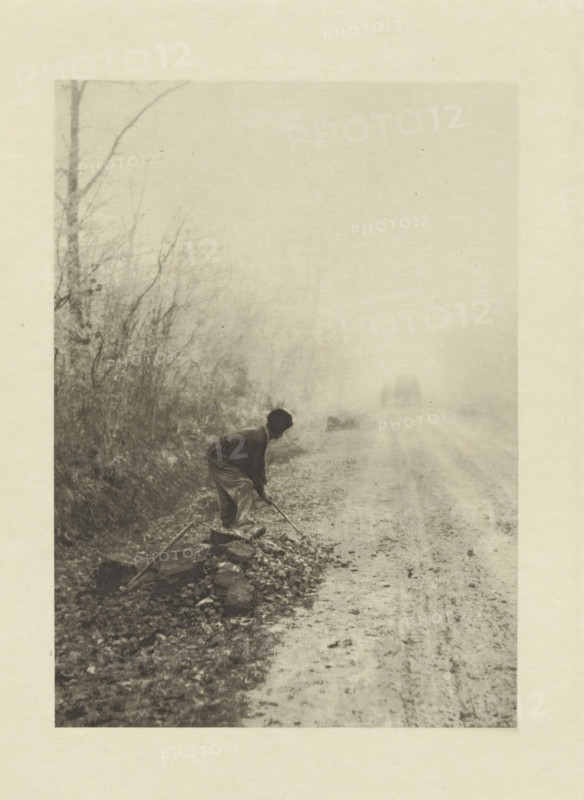 Worker along the road, Dans le brouillard, photomechanical print, photographer: Jean-Ferdinand Coste, cliché maker: Fillon & Heuse, printer (person): Charles Wittmann, Paris, c. 1891 - in or before 1896, printer (person), paper, photoaquatint, height 165 mm x width 118 mm, road, path, working class, laborers, A grainy monochrome scene shows a person bending forward near the edge of a rough road, holding a long tool and working among scattered stones and debris, with a line of bare trees and tangled underbrush along the roadside fading into a misty background and a faint silhouette of a horse-drawn vehicle visible down the road.
