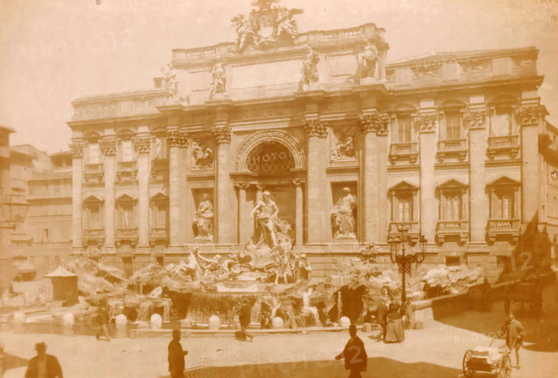 Roma, Fontana di Trevi, Trevi Fountain in Rome, photograph, page, number: '1014 ?', Rome, c. 1883 - in or before 1888, photographer, paper, albumen print, height 100 mm x width 140 mm, Photo on page 120., ornamental fountain, Trevi Fountain, A faded photograph of a grand stone building with ornate columns and sculpted figures on its façade, a large arched central window above a prominent sculpted fountain with flowing water and multiple statues, rocky basins at the fountain's base, several silhouetted people and a person pulling a small cart in the foreground, surrounding buildings partially visible at the sides, and an overall warm, sepia-toned appearance.