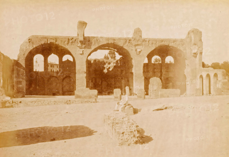 Ruin of the Temple of Peace in Rome, Roma, Foro Romano - tempio della pace, photograph, page, number: '1036.', Rome, c. 1883 - in or before 1888, photographer, paper, albumen print, height 100 mm x width 140 mm, Photo on page 137., landscape with ruins, Peace Temple, A faded photograph of a large stone structure with a series of tall rounded arches set on a raised platform, showing weathered surfaces and areas of erosion, with smaller archways visible within the larger arches and a few freestanding stone blocks and rubble scattered in front; a figure seated on a low stone in the foreground, indistinct in detail, and a patch of darker ground casting a shadow at the lower left, with a pale sky above.