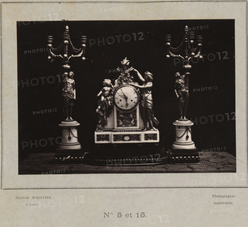 Clock and two candlesticks, photograph, page, inscription: 'Photographie, inaltérable.', number: 'PL. III., Nos 5 et 15.', photographer: Paul Joseph Nicolas Nestor Schaffers, 1873 - 1878, paper, carbon print, height 95 mm x width 134 mm, chandelier, candelabrum, clock, A black rectangular photograph mounted on a pale mat shows an ornate clock set flanked by matching candelabra on a patterned surface; the central clock features a round face with hands, sculptural floral and ribbon decorations, a reclining cherub figure on one side, and a draped female figure leaning against the clock on the other side, while each candelabrum rises from a columnar base topped with a standing draped female figure and branching arms with decorative chains and bulbous ornaments.