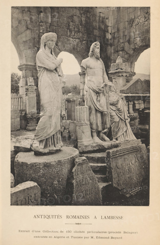 Antiquités romaines à Lambesse, Roman antiquities at Lambaesis, page, photomechanical print, inscription: 'Phototypie Berthaud - Paris, Extrait d'une Collection de 150 clichés pelliculaires (process Balagny), exécutes en Algérie et Tunisie par M, Edmond Bayard', photographer: Edmond Bayard, Lambaesis, cliché maker: Michel Berthaud, Paris, c. 1881 - in or before 1886, Edmond Bayard, Michel Berthaud, photographer, cliché maker, North Africa, paper, collotype, photoprint, height 165 mm x width 116 mm, piece of sculpture, reproduction of a piece of sculpture, statues, paintings, etc., objects of worship in Roman religion, inscription, The image shows a group of weathered stone statues and architectural fragments arranged among ancient ruins. In the foreground, a draped female figure stands on a round pedestal with folded arms and a covered head, her garments falling in vertical folds to her feet. Beside her, a bearded male figure with a partially draped torso stands on a separate base, his...