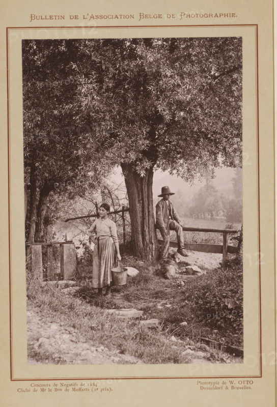 Un coin du Condroz (original title), Boy and a woman by a fence under a tree, Condroz, page, photomechanical print, inscription: 'Concours de Mr le Bon de Moffarts (Ir prix).', photographer: Armand baron de Moffarts, clichémaker: Wilhelm Otto, Brussels, c. 1879 - in or before 1884, printer (person), pasteboard, paper, collotype, photogravure, height 148 mm x width 98 mm, Print on unnumbered page., boy (child between toddler and youth), fence, wall, paling, farmers - AA - female farmer, farmer's wife, trees, A sepia-toned photograph mounted on a page shows a large tree with dense foliage dominating the upper portion and center. Beneath the tree, a young woman in a long skirt and blouse stands holding a metal pail at her side, looking slightly to her left. Nearby, a man wearing a jacket and wide-brimmed hat sits on a wooden rail fence with one leg bent and the other hanging down, facing forward. A dog lies on the ground near the man's feet. Grass, scattered stones, and small plants cover...