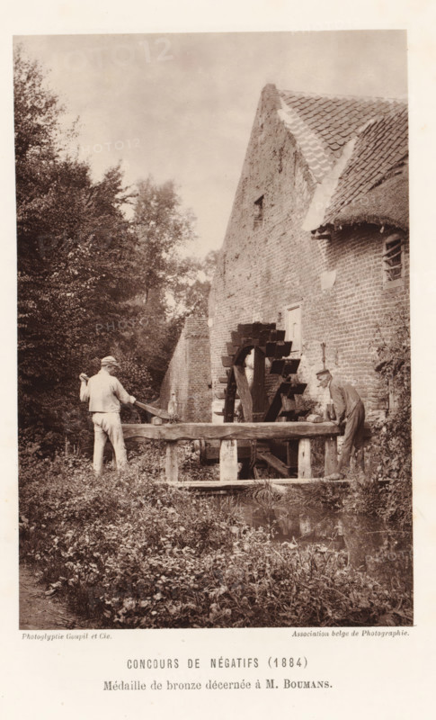 Two men adjust the water level at a watermill, in Eijsden, page, photomechanical print, inscription: 'Concours de négatifs (1884), Médaille de bronze décernée à M. Boumans.', photographer: Mathias Boumans, Eijsden, clichémaker: Goupil & Cie., Paris, c. 1879 - in or before 1884, printer (person), paper, Woodburytype, Woodburytypie, height 149 mm x width 98 mm, Print on unnumbered page., watermill, water mill, A sepia-toned vertical photograph shows an old brick building with a steep tiled roof and a thatched section, a wooden waterwheel attached to the building, and a wooden trough structure in front of the wheel. Two men stand by the trough facing the wheel; one man on the left wears light clothing and a cap and holds a long tool over his shoulder, the other man on the right leans forward wearing darker clothing and a hat. Dense foliage and trees occupy the left side and background, and grassy plants and low vegetation fill the foreground with a small reflective water surface near the ...